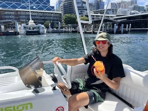 A guest at Capri's helm with a drink, Darling Harbour's wharves behind