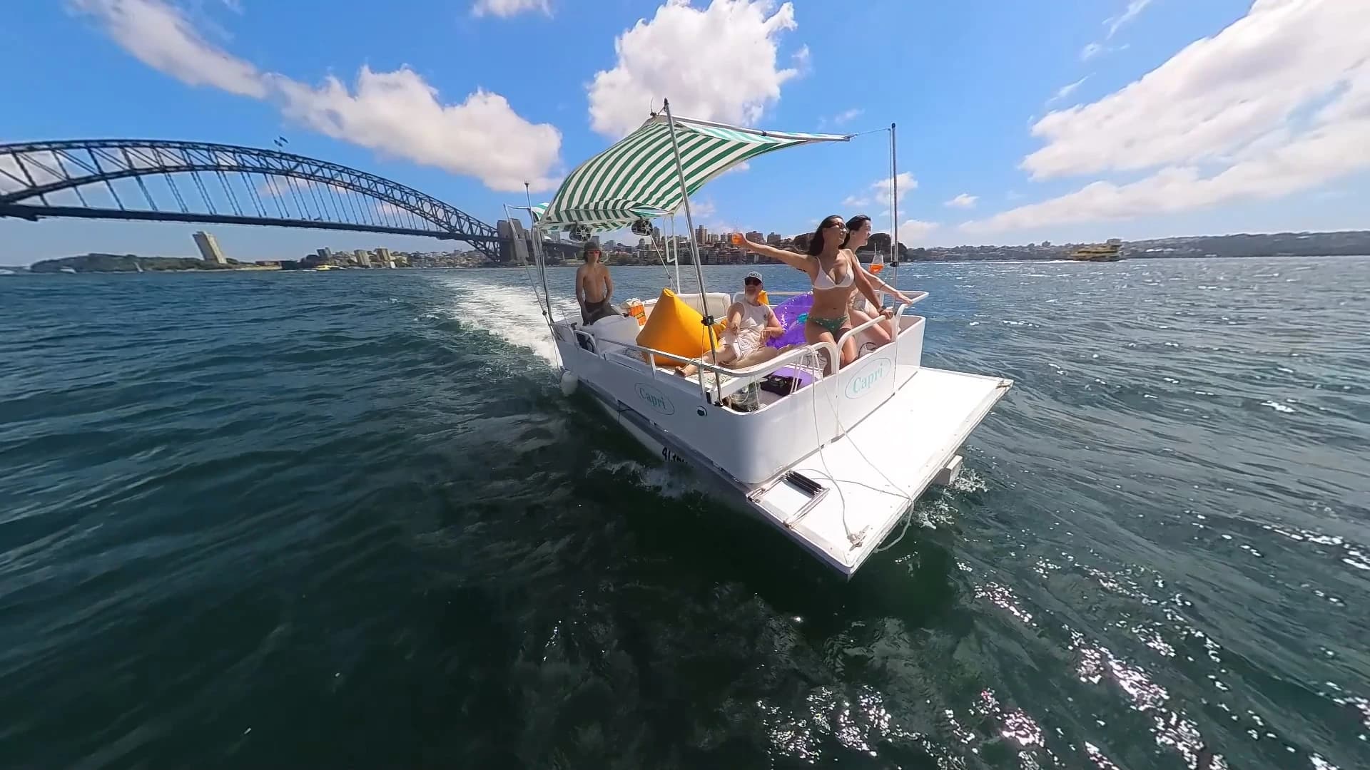 Capri underway on Sydney Harbour with the Harbour Bridge behind, four guests under the striped canopy and the yellow beanbag up front